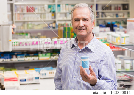 Senior male pharmacist holding medicine container at pharmacy counter with shelves and dispenser Senior male pharmacist holding medicine container at pharmacy counter with shelves and dispenser 133290836