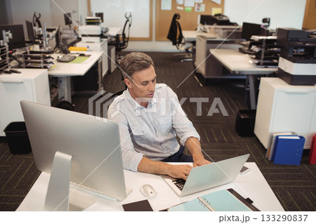 Middle-aged man in business casual attire typing on laptop at desk in open-plan office with monitor Middle-aged man in business casual attire typing on laptop at desk in open-plan office with monitor 133290837