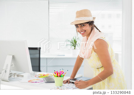 Woman in twenties leaning over office desk wearing straw fedora hat reviewing color swatches 133290848