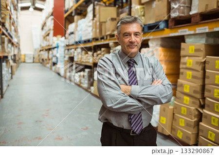 Mature adult man standing with arms crossed in warehouse aisle among shelving units, copy space 133290867
