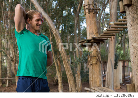 Mid adult man stretching triceps in green shirt at ropes course amid rope bridges, copy space 133290868