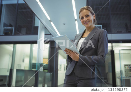 Businesswoman in suit holding tablet, looking at camera in office with glass partitions, copy space 133290875