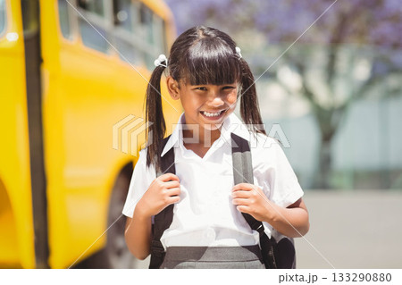 Child girl in school uniform standing on pavement by yellow school bus, carrying black backpack Child girl in school uniform standing on pavement by yellow school bus, carrying black backpack 133290880