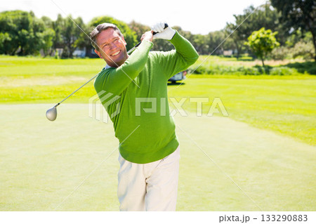Senior male golfer swinging driver above shoulder on putting green with golf cart and glove 133290883