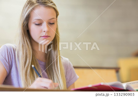 Teenage girl in lilac t-shirt writing notes in notebook at classroom desk with pencil, copy space Teenage girl in lilac t-shirt writing notes in notebook at classroom desk with pencil, copy space 133290903