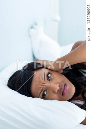 African American woman lying on white pillow on bed in bedroom pressing hand against ear 133290908