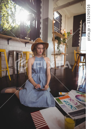 Woman kneeling on dark floor in bright cafe using smartphone amid design sheets and color swatches 133290916