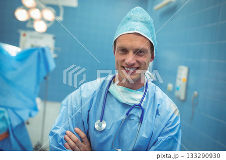 Mid adult male surgeon wearing scrubs crossing arms by OR table under OR lights with stethoscope Mid adult male surgeon wearing scrubs crossing arms by OR table under OR lights with stethoscope 133290930