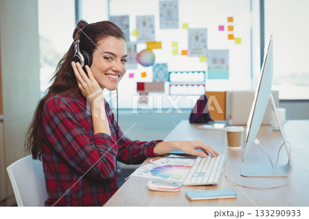 Female graphic designer smiling while adjusting headphones at studio desk with color wheel printout Female graphic designer smiling while adjusting headphones at studio desk with color wheel printout 133290933