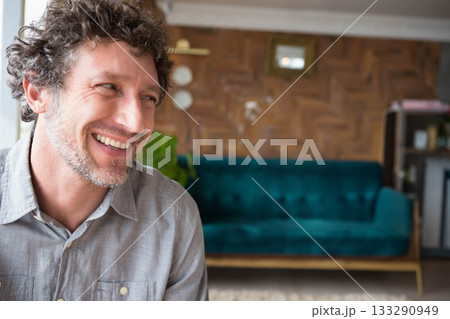 Middle-aged man smiling and sitting in living room with teal sofa, potted plant and shelving unit Middle-aged man smiling and sitting in living room with teal sofa, potted plant and shelving unit 133290949