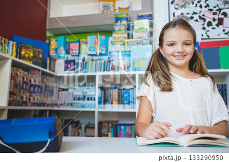 Female child sitting at library desk reading open book with white shelves, blue device, copy space Female child sitting at library desk reading open book with white shelves, blue device, copy space 133290950
