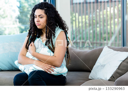 Woman in her twenties hugging decorative pillow, sitting on sofa by vertical blinds, copy space 133290963