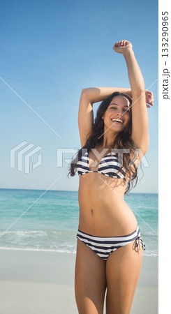 Woman standing on sandy beach raising arms under clear blue sky wearing striped bikini, copy space Woman standing on sandy beach raising arms under clear blue sky wearing striped bikini, copy space 133290965
