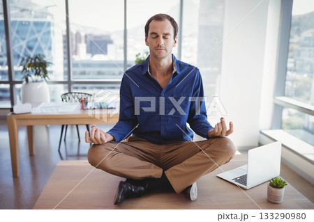 Mid-adult man meditating cross-legged on wooden desk in open-plan office with laptop and plants Mid-adult man meditating cross-legged on wooden desk in open-plan office with laptop and plants 133290980