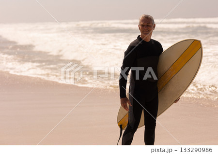 Senior man standing knee-deep at beach holding surfboard and wearing wetsuit, copy space Senior man standing knee-deep at beach holding surfboard and wearing wetsuit, copy space 133290986