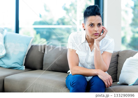 Woman wearing shirt and trousers sitting on taupe sofa with pillows in living room by windows Woman wearing shirt and trousers sitting on taupe sofa with pillows in living room by windows 133290996