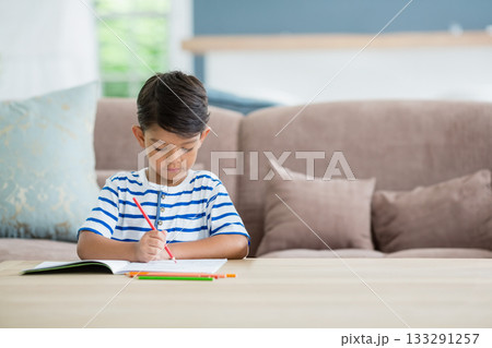 Asian boy holding red pencil while coloring notebook at wooden coffee table in living room 133291257