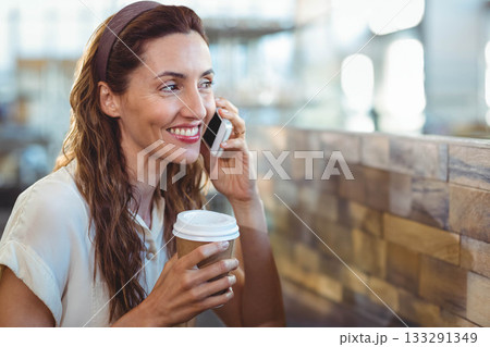 Middle-aged woman smiling while holding paper coffee cup and talking on smartphone at cafe counter 133291349