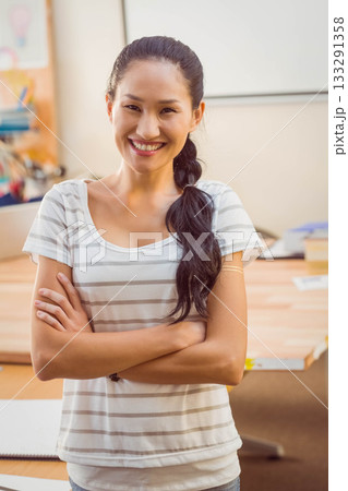 Asian woman standing crossing arms and smiling at classroom desk with whiteboard and art supplies Asian woman standing crossing arms and smiling at classroom desk with whiteboard and art supplies 133291358