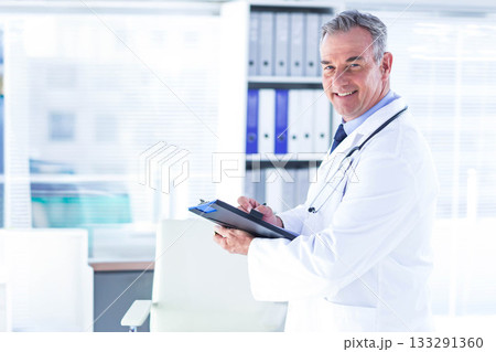 Male doctor writing clipboard with pen in lab coat stethoscope at clinic by shelving, copy space Male doctor writing clipboard with pen in lab coat stethoscope at clinic by shelving, copy space 133291360