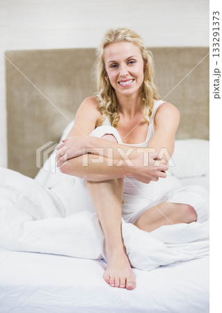 Woman wearing pajamas sitting on bed hugging knee and smiling in bedroom with pillows and headboard 133291373