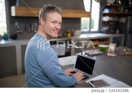 Mature male sitting at kitchen island, working on laptop with tablet, documents and mixing bowl 133291386