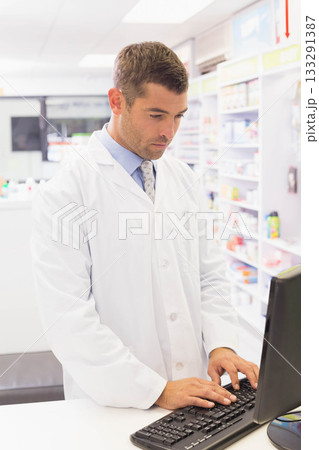 Mid adult male pharmacist typing on keyboard behind counter at pharmacy with medication shelves 133291387