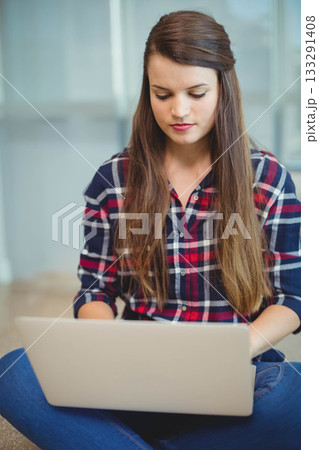 Woman sitting cross-legged on floor working on silver laptop in modern office with glass panels Woman sitting cross-legged on floor working on silver laptop in modern office with glass panels 133291408