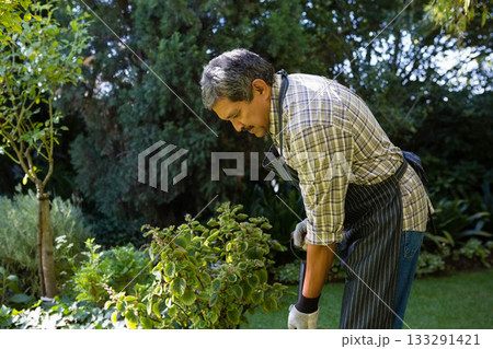 Senior man leaning over shrub in backyard garden using trowel with apron and gloves, copy space Senior man leaning over shrub in backyard garden using trowel with apron and gloves, copy space 133291421