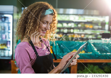 Woman in black apron tapping tablet at produce bins near refrigerated display case, copy space 133291430