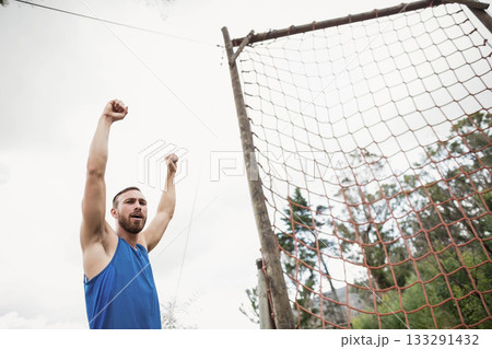 Man raising both arms overhead beside rope net obstacle at wooded clearing with wooden poles Man raising both arms overhead beside rope net obstacle at wooded clearing with wooden poles 133291432