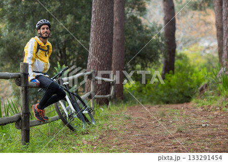Man sitting on split-rail fence beside mountain bike on trail wearing helmet, daypack, copy space 133291454