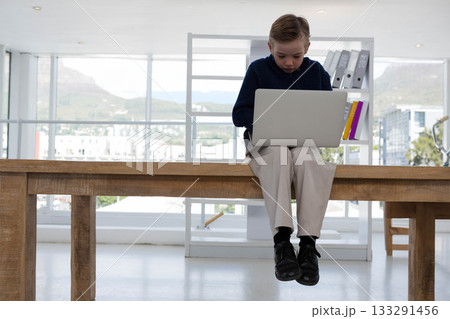 Boy child sitting on edge of table in bright study using silver laptop near ring binders 133291456