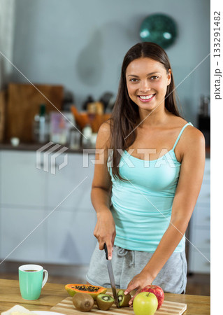Woman slicing kiwis papaya apples with chef's knife on wooden countertop in kitchen with green mug 133291482