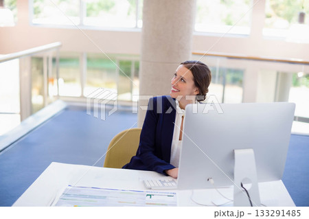 Female office worker wearing navy blazer smiling, looking up at monitor at office desk, copy space Female office worker wearing navy blazer smiling, looking up at monitor at office desk, copy space 133291485