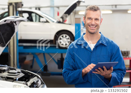 Mid-adult man wearing coveralls holding tablet in bay near open hood with lifted sedan, copy space Mid-adult man wearing coveralls holding tablet in bay near open hood with lifted sedan, copy space 133291502