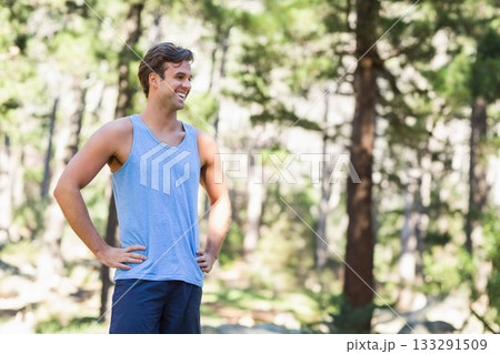 Man standing with hands on hips smiling in pine forest clearing among rocks under dappled sunlight Man standing with hands on hips smiling in pine forest clearing among rocks under dappled sunlight 133291509