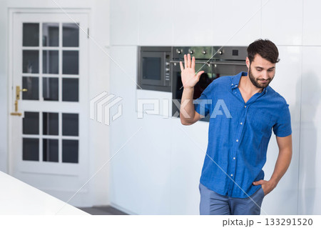 Man in late twenties standing in kitchen by integrated appliances and glass door waving, copy space 133291520