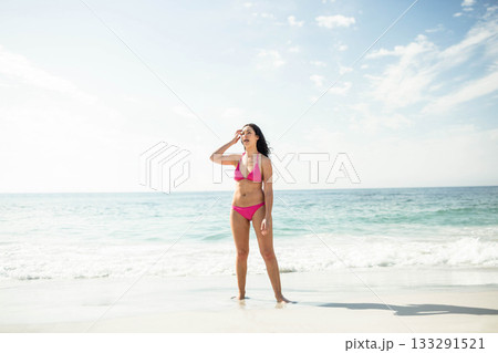 Woman standing barefoot at beach edge wearing pink bikini, shielding eyes and gazing toward horizon Woman standing barefoot at beach edge wearing pink bikini, shielding eyes and gazing toward horizon 133291521