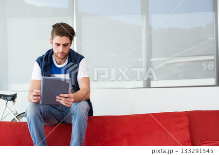 Man using tablet computer while sitting on red couch in bright lounge area with bicycle frame 133291545