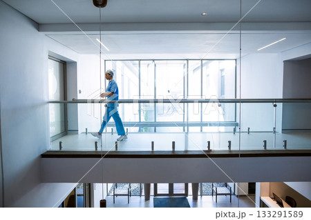 Asian woman in scrubs walking along hospital hallway with glass railing and windows, copy space 133291589
