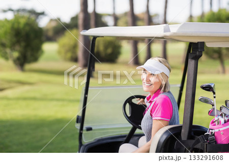 Female golfer gripping steering wheel while sitting in golf cart on sunlit fairway with golf bag 133291608