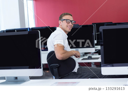 Male student smiling while sitting at desk in computer lab using computer monitors and keyboard 133291613