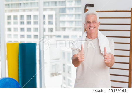 Senior man standing in fitness studio giving thumbs up with towel draped, stall bars, copy space 133291614