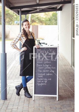 Female server standing under café walkway by bicycle, leaning on menu board, copy space 133291653