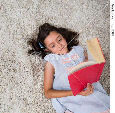 Child girl wearing light blue blouse reading hardcover red book on cream-colored shag rug at home 133291660