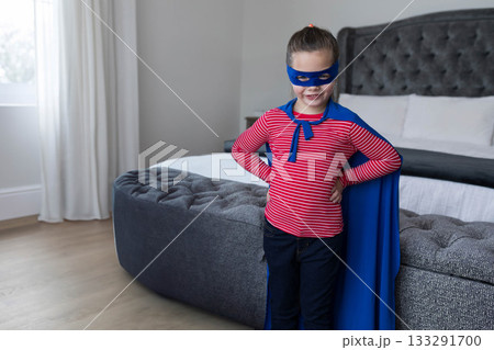 Child girl wearing blue cape, mask, striking pose beside bench at foot of bed, copy space Child girl wearing blue cape, mask, striking pose beside bench at foot of bed, copy space 133291700
