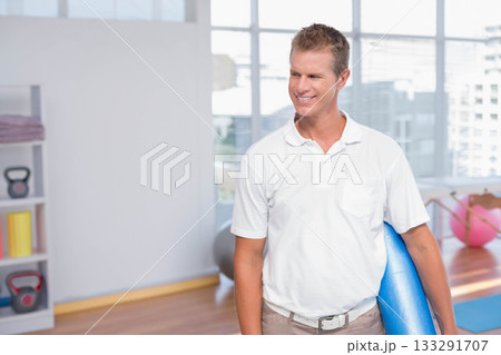 Man standing in fitness studio wearing polo shirt holding blue yoga mat over shoulder, copy space Man standing in fitness studio wearing polo shirt holding blue yoga mat over shoulder, copy space 133291707
