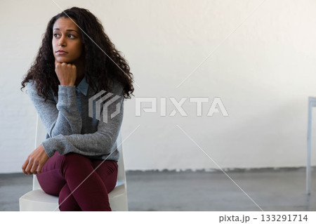 Woman sitting pensively on white chair in studio wearing grey sweater with table leg, copy space 133291714