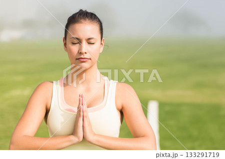 Woman standing in grassy field wearing light-colored sports top pressing hands together in prayer Woman standing in grassy field wearing light-colored sports top pressing hands together in prayer 133291719